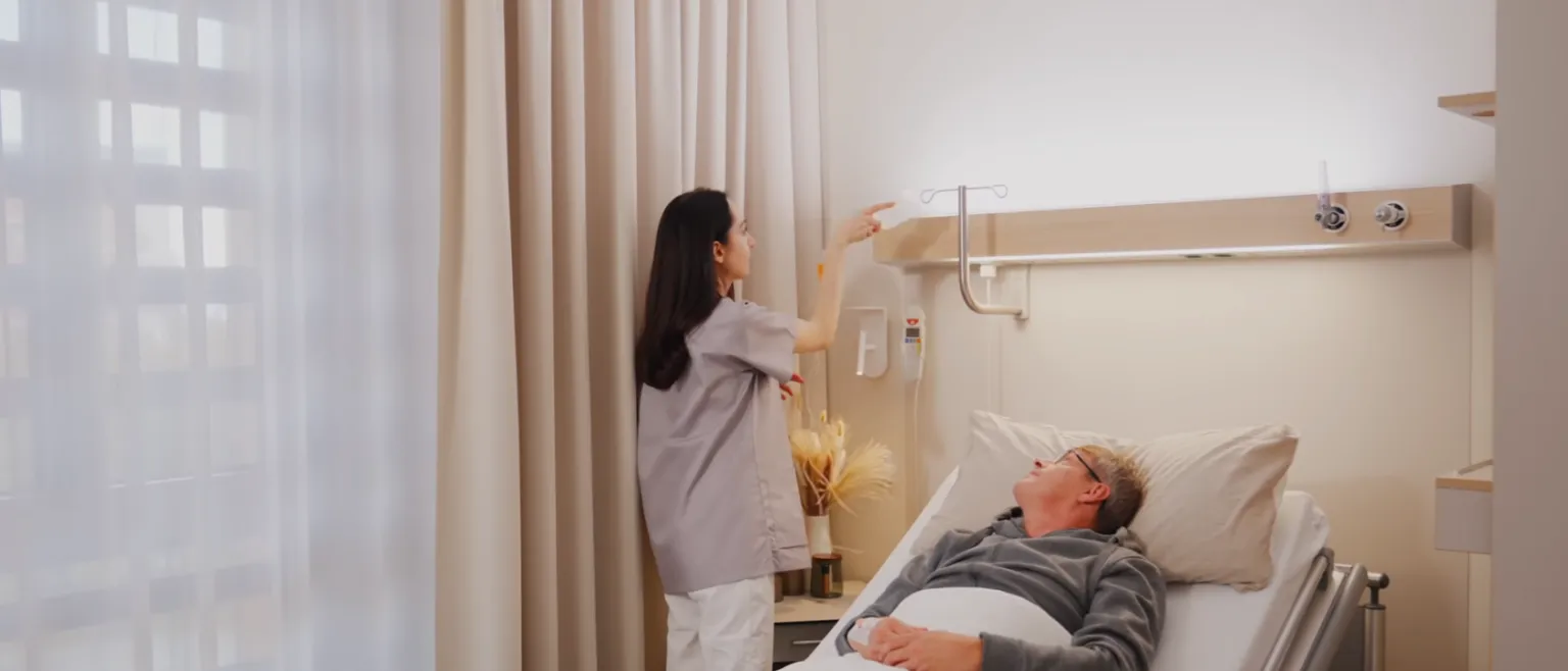 Krankenpflegekraft steht neben einem liegenden Mann in einem Krankenhausbett und greift nach einem Infusionsständer an der Wand