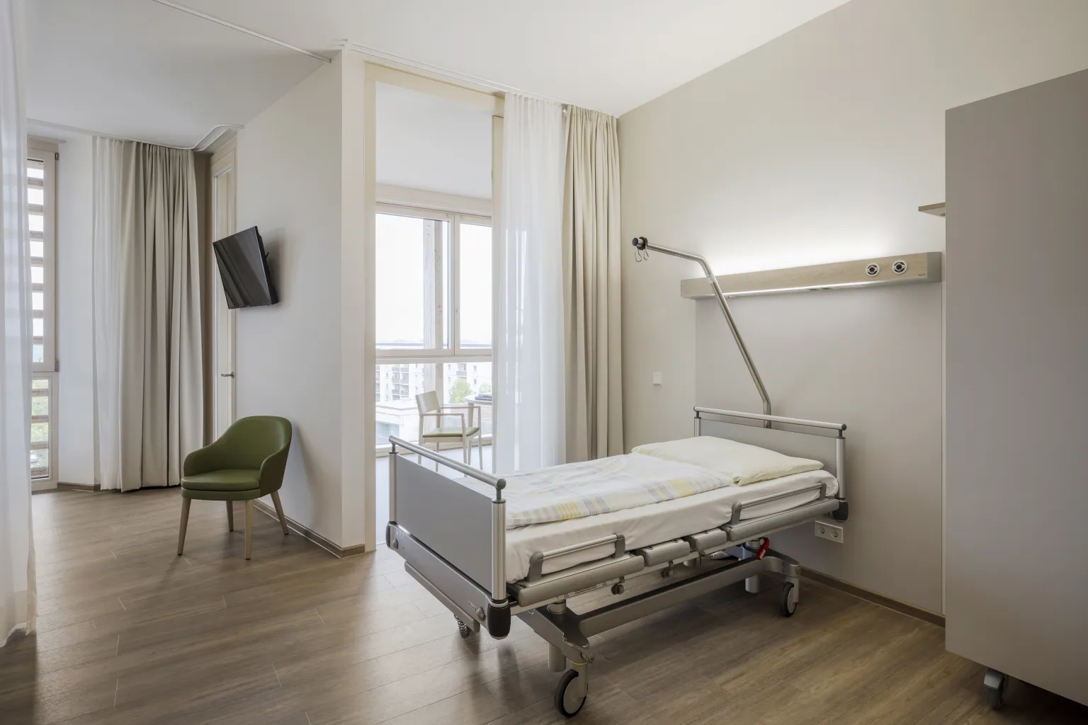 Krankenhauszimmer mit einem Bett, grünem Stuhl, Fenster mit Vorhängen und Fernseher an der Wand