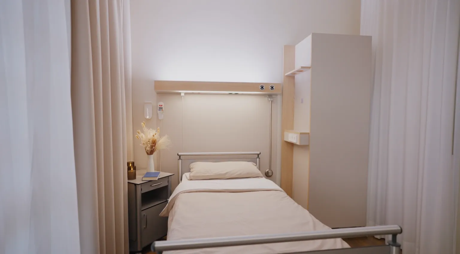 Ein einzelnes Krankenhausbett mit weißer Bettwäsche steht in einem hellen Raum. Links neben dem Bett befindet sich ein kleiner Nachttisch mit einer Vase, die getrocknete Gräser enthält, und einem geschlossenen Buch. Die Wände und Vorhänge sind in hellen, neutralen Tönen gehalten. Über dem Bett ist eine Leuchte an der Wand angebracht, die den Bereich ausleuchtet. Rechts steht ein schlichter, heller Schrank mit offenen Fächern.