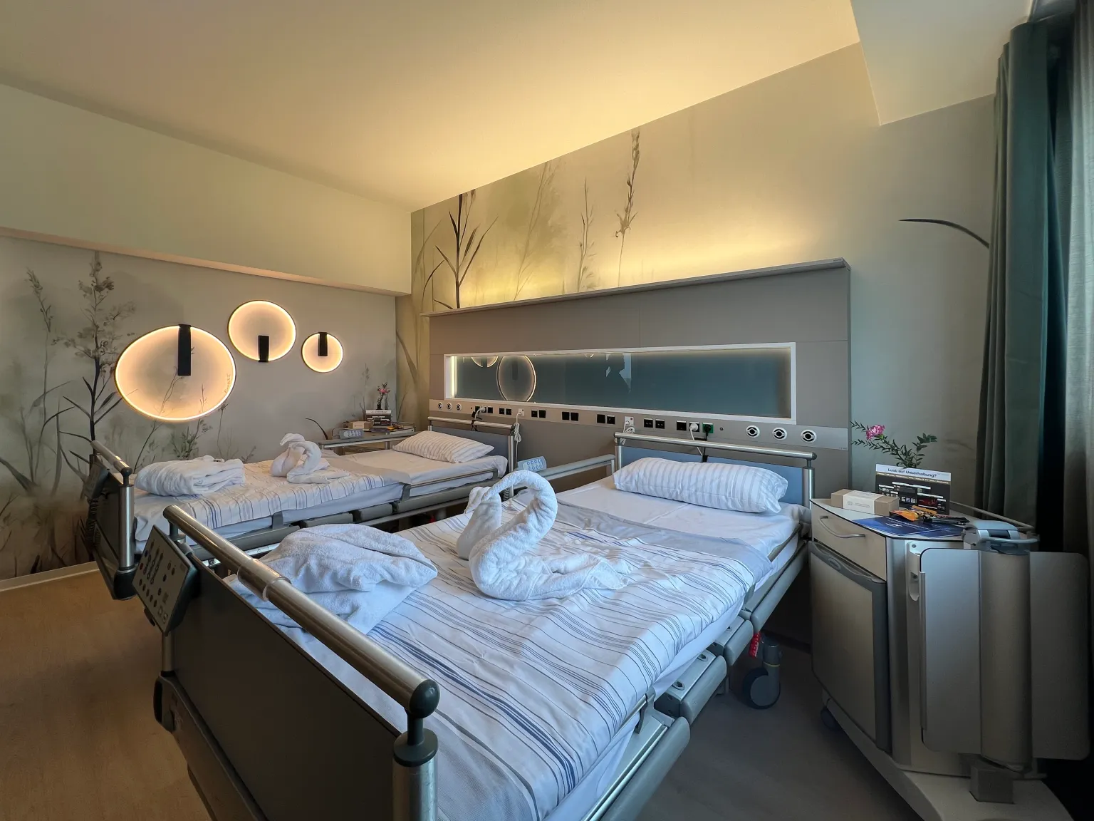Zwei Krankenhausbetten mit weißen Bettlaken und Handtüchern, die zu Schwänen gefaltet sind, in einem Zimmer mit indirekter Beleuchtung und Wanddekoration mit Pflanzenmotiven